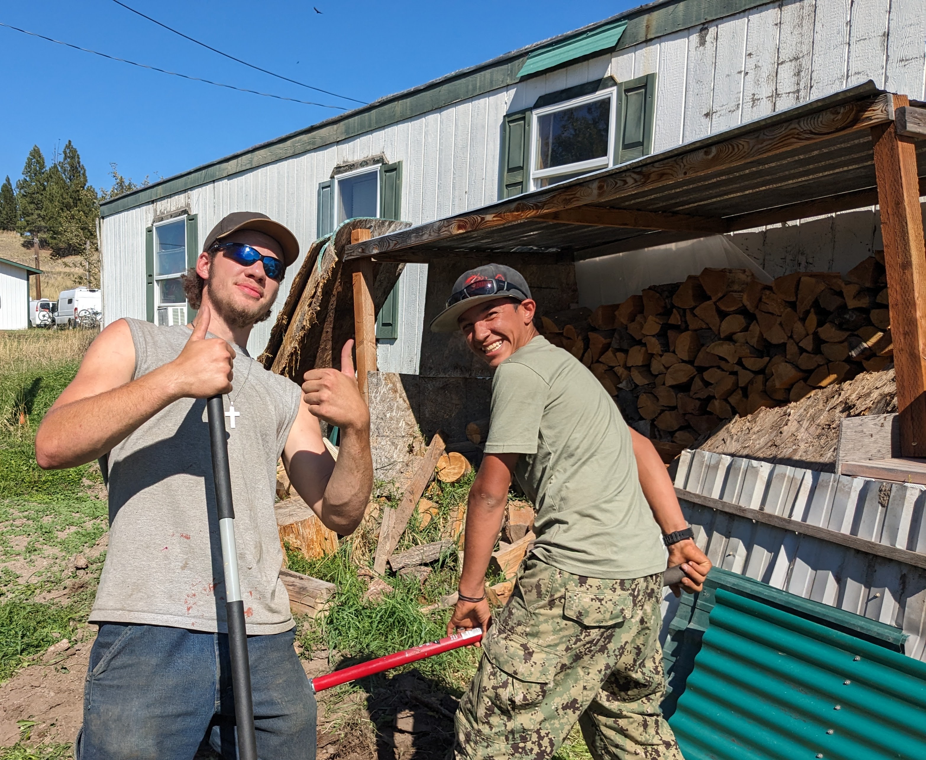 Two young volunteers grinning with thumbs up in front of a stacked woodshed